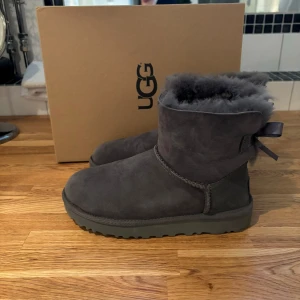 Grå UGG boots med rosett - UGG bailey bow - supermysiga grå UGG boots i mocka med en söt rosett på baksidan. De är i ny  skick, endast använda ett fåtal gånger inne och kommer i originalkartong. Nypris 2700 säljes för 1000:- Kan skickas eller hämtas i Täby C 