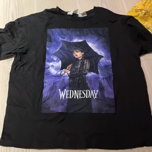 Svart t-shirt med Wednesday-motiv från H&M - Cool svart t-shirt från H&M med ett tryck av Wednesday i lila och blått. Perfekt för fans av den mystiska stilen! T-shirten är kortärmad och gjord i mjuk bomull, vilket gör den bekväm att bära hela dagen.