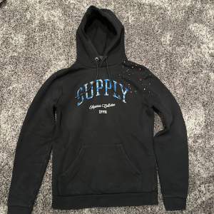 Säljer en svart hoodie med texten 'SUPPLY' i blått och vitt på bröstet. Den har en cool färgstänkseffekt i rött, vitt och blått på ena axeln. Perfekt för en avslappnad stil. Den har en klassisk känguruficka och justerbar huva.