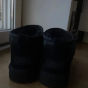 Svarta UGG mini i mocka - Säljer ett par svarta UGG i mocka. De är i bra skick och perfekta för höst och vinter. Skorna har en enkel design med en liten dragflik baktill för lättare påtagning. De är varma och bekväma, idealiska för kyligare dagar.