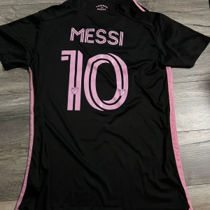 Svart och rosa fotbollströja - Säljer en snygg svart fotbollströja med rosa detaljer från Adidas. Tröjan har korta ärmar och Messi 10 tryckt på ryggen. Perfekt för fotbollsfans som vill ha en stilren look på planen eller på stan. Materialet är lätt och andas bra. Passar både till träning och vardag.