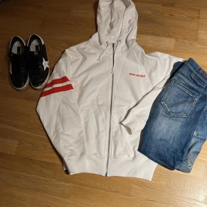 Vit hoodie från Helmut Lang - Säljer en snygg vit hoodie från Helmut Lang med röda detaljer. Den har en dragkedja framtill och texten 'TEAM HELMUT' på bröstet. På ryggen står det 'HELMUT LANG' i rött. Skicket är som ny. Aldrig använd och köpt från sicuro fashion. Storlek S