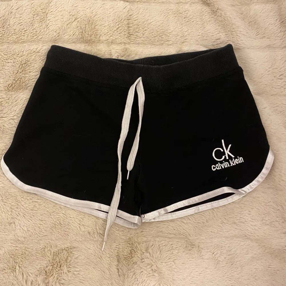 Snygga svarta shorts från Calvin Klein med vit kant och logga. De har en bekväm passform med snörning i midjan. Perfekta för en avslappnad dag eller träning. 🖤. Shorts.