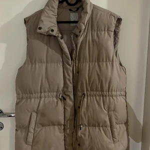 Beige dunväst från Etrel - Säljer en snygg beige dunväst från Etrel i storlek 40. Västen har en dragkedja och knappar framtill, perfekt för höst och vinter. Den är i mycket bra skick och supermysig att ha på sig när det är kyligt ute. Passar både till vardags och lite mer avslappnade tillfällen.