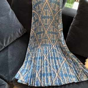 Blå mönstrad klänning från Sandro - Säljer en superfin blå kjol från Sandro med ett unikt mönster i vitt och svart. Klänningen har en elegant plisserad design som ger en snygg rörelse. Perfekt för både vardag och fest! 💙