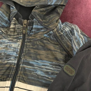  jacka från Boss - Säljer en cool jacka från Boss med ett unikt mönster i blått och grått. Jackan har en dragkedja framtill och en huva för extra stil och komfort. Perfekt för höst och vår, med långa ärmar och en normal passform. Den är i mycket bra skick och redo för nya äventyr!