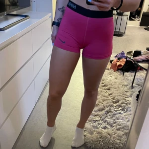 Rosa träningsshorts från Nike - Säljer ett par snygga rosa träningsshorts från Nike Pro. De har en tight passform som är perfekt för träning. Shortsen har en svart resårmidja med Nike-loggan och är gjorda i ett bekvämt och stretchigt material.