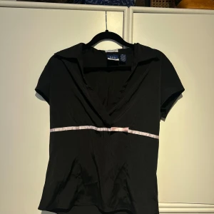 Svart topp med rosett  - Snygg svart topp med en söt liten rosett i midjan under bysten. Toppen har en v-ringad design och korta ärmar, vilket gör den perfekt för både vardag och fest. Materialet är stretchigt och bekvämt, vilket ger en bra passform.