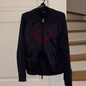 Mörkblå Armani Cardigan  - Snygg mörkblå cardigan ish från Armani Jeans med röd logga på bröstet. Jackan har dragkedja och är långärmad, perfekt för höst och vår. Materialet är en mix av syntet och bomull vilket gör den både bekväm och stilren. Passar perfekt för en casual look eller en utekväll. Den har en liten slitning vid bröstet vilket man kan se på bilderna. Den är M men passar även storlek S. 