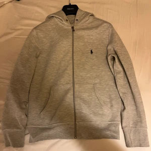 Grå zip hoodie Ralph Lauren - Säljer en stilren grå hoodie från Ralph Lauren i bra skick. Den har en dragkedja framtill och en klassisk logga broderad på bröstet. Perfekt för både vardag och chill dagar. Storleken är S. Passar från 175-183