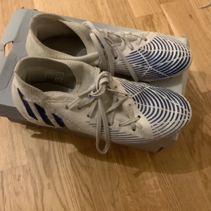 Vita och blå fotbollsskor från Adidas - Säljer ett par vita och blå fotbollsskor från Adidas i mycket bra skick. Skorna har ett coolt mönster med blå ränder och snörning för perfekt passform. Perfekta för fotbollsträning eller match!