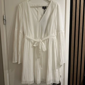 Vit klänning från Bubbleroom - Säljer en elegant vit klänning från Bubbleroom i storlek 42. Helt oanvänd, endast testad. Klänningen har en v-ringad design med långa ärmar och ett knytband i midjan. Den är tillverkad i 100% polyester och har ett plisserat mönster på kjolen och ärmarna. Perfekt för sommarens fester eller en stilren look på våren.