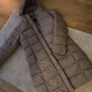 Beige dunjacka med huva - Säljer en supermysig beige dunjacka med huva. Perfekt för kalla vinterdagar! Jackan är långärmad och har en quiltad design som håller dig varm och stilren. Den är i mycket bra skick och passar perfekt för både vardag och vinteräventyr. Storlek XS 34