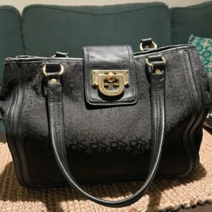 Svart handväska från DKNY - Snygg svart handväska från DKNY med ett monogrammönster. Väskan har guldfärgade detaljer och en stilren låsdetalj framtill. Perfekt för både vardag och fest! 🖤