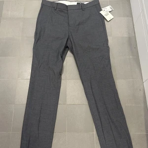 Grå kostymbyxor nyskick - Säljer ett par stiliga grå kostymbyxor från H&M i storlek 48. De är i nytt skick och ger en modern look. Perfekta för både kontoret och festliga tillfällen. Materialet är en blandning av polyester, viskos och bomull vilket gör dem bekväma och hållbara.