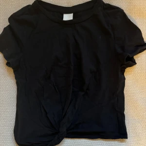 Svart kortärmad topp från H&M - Säljer en enkel och stilren svart kortärmad topp från H&M i storlek XS. Perfekt för vardagsbruk eller att styla upp för en kväll ute. Toppen har en liten knutdetalj framtill som ger en extra touch. Passar bra till jeans eller kjol!