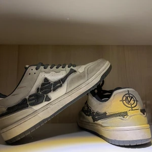 Beige och svarta sneakers från Vicinity - Säljer ett par coola sneakers från Vicinity i beige och svart med unikt mönster på sidorna. Skorna har svarta snören och en robust sula, perfekt för vardagsbruk. De är i bra skick med lite slitage på sulan. Perfekta för höst och vår!