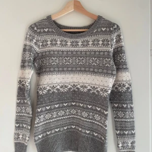 Grå stickad tröja från H&M - Mysig grå stickad tröja från H&M med ett klassiskt mönster i vitt. Perfekt för kalla dagar med sina långa ärmar och ribbade muddar. Tröjan har en rund halsringning och är i mycket bra skick. Passar perfekt för både höst och vinter!
