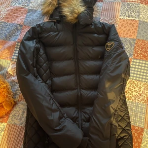 Svart dunjacka från Vinson Polo Club - Snygg svart dunjacka från Vinson Polo Club med quiltade detaljer och en mysig pälskantad huva. Jackan har en dragkedja framtill och ett stilrent märke på ärmen. Perfekt för kyliga dagar.