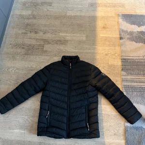 Svart pufferjacka - Snygg svart pufferjacka med dragkedja och hög krage. Perfekt för kyliga dagar med sin quiltade design och praktiska fickor med dragkedjor.