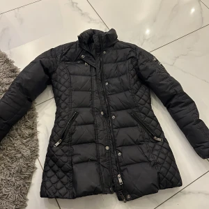 Svart quiltad jacka från Rockandblue - Snygg svart quiltad jacka från Rockandblue med dragkedja och knappar. Jackan har en hög krage och långa ärmar, perfekt för kyligare dagar. Den har praktiska fickor med dragkedjor och ett stilrent märke på ärmen.