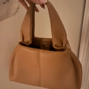 Brun/orange handväska  - Väskan har två  handtag och en knappstängning upptill, tillkommer även ett längre band man kan fästa på sidorna. Jättesöt och typ aldrig använd