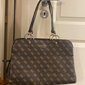 Mönstrad axelväska från Guess - Snygg axelväska från Guess med ett elegant mönster i brunt och beige. Perfekt för att hålla dina saker organiserade och stiliga. Defekter: Sönder dragkedja men går nog att fixa.