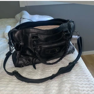 Svart axelväska med dragkedja - Snygg svart axelväska i skinn med flätade handtag och justerbar axelrem. Väskan har flera praktiska fack och en dragkedja för säker förvaring. Perfekt för att bära med sig det nödvändigaste med stil.