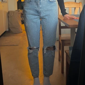 Blå jeans med slitningar - Säljer ett par klassiska blå jeans (perfect jeans)  från gina med hög/medel midja och slitningar på knäna. De har en straight passform och är perfekta för en avslappnad stil. Jeansen har fyra fickor och en knappgylf.