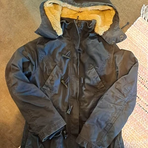 Ljussvart parkajacka med teddyfoder - Säljer en ljussvart parkajacka med mysigt teddyfoder och huva. Jackan har knappar och dragkedja framtill samt praktiska fickor. Perfekt för kyliga dagar!