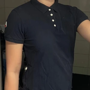 Mörkblå pikétröja med knappdetaljer - Snygg mörkblå pikétröja med korta ärmar och klassisk krage. Tröjan har vita knappar framtill och en logotyp på ärmen. Perfekt för en stilren och avslappnad look. Från CRW och hööks 