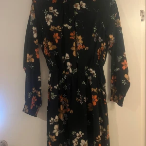 Blommig långklänning med hög krage - Snygg långklänning med blommönster i orange, vitt och grönt på en svart bakgrund. Klänningen har en hög krage med knäppning och långa ärmar. Perfekt för höst och vår.