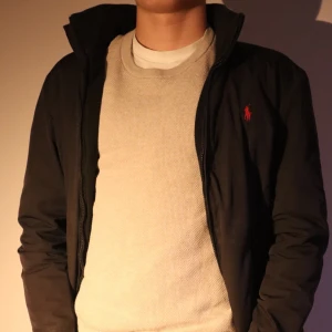 Svart jacka från Polo Ralph Lauren - Säljer en stilren svart jacka från Polo Ralph Lauren med det klassiska röda logotypbroderiet på bröstet. Jackan har en enkel design med dragkedja och är perfekt för en avslappnad look. Tecken på användning