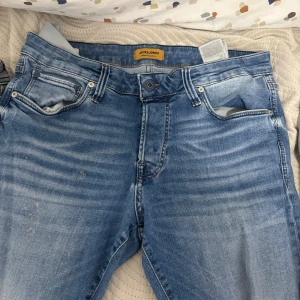 Blå jeans från Jack & Jones - Snygga blå jeans från Jack & Jones i modellen Slim/Glenn. De har en klassisk femficksdesign med knappgylf och är perfekta för en stilren look. Passar bra till både vardag och mer uppklädda tillfällen.