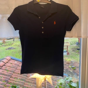Svart pikétröja från Ralph Lauren - Säljer en stilren mörkblå pikétröja från Ralph Lauren med orange logga på bröstet. Tröjan har korta ärmar och en klassisk krage med knappar. Perfekt för en avslappnad men ändå elegant look.