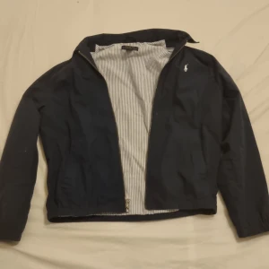 Mörkblå jacka från Polo Ralph Lauren - Säljer en stilren mörkblå jacka från Polo Ralph Lauren. Jackan har en dragkedja framtill och ett diskret broderat märke på bröstet. Insidan är fodrad med ett randigt mönster. Perfekt för en klassisk look. Är 160 cm eller xs i storlek