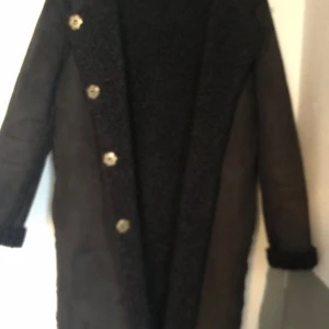 Svart kappa med knappar - Elegant svart kappa med långa ärmar och knappar framtill. Den har en mysig, pälsliknande insida och detaljer längs kanterna. Perfekt för kyliga dagar.