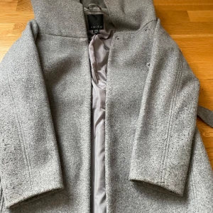 BUDA PÅ:Grå kappa från Amisu - Elegant grå kappa från Amisu i ullblandning. Den har en stilren design med knappar och ett bälte i midjan för en snygg passform. Perfekt för kyliga höst- och vinterdagar.