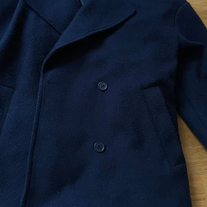 BUDA: Mörkblå kappa från NELLY  - Oversized vibe; Elegant mörkblå kappa från NELLY med klassisk krage och knappar framtill. Perfekt för kyligare dagar med sin stilrena design och långa ärmar. Ett måste i garderoben för en tidlös look.