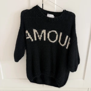 Svart stickad tröja från Noella - Säljer en svart stickad tröja från Noella med texten 'AMOUR' i vitt på framsidan. Tröjan har trekvartsärmar och en avslappnad passform. Perfekt för en mysig dag!