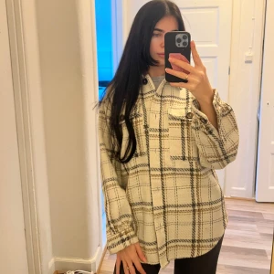 Rutig overshirt i beige och svart - Snygg och trendig overshirt i beige och svart rutmönster. Den har långa ärmar och knappar framtill. Perfekt för lager-på-lager-stilen. Storlek S.