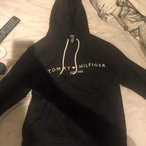Svart hoodie från Tommy Hilfiger - Snygg svart hoodie från Tommy Hilfiger med vit logotyp på bröstet och justerbar huva med vita snören. Perfekt för en avslappnad stil. Använd 3 gånger Max men då jag är i behov av pengar säljer jag den. Köpt på Zalando och nypris är 1200kr. Säljes enbart med swish och köparen står för frakten.