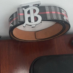 Burberry bälte - Snyggt rutig bälte från Burberry med ett stort silverfärgat spänne. Bältet har ett klassiskt mönster i svart, grått och rött med vita detaljer. Perfekt för att ge din outfit en stilren touch. Säljer för behöver mer pengar använd bara ett par gånger. Kom dm för mer bilder