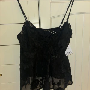 Svart blommig topp från Gina Tricot - Snygg svart topp från Gina Tricot med blommigt mönster och tunna axelband. Toppen har en knytning framtill och är tillverkad i ett lätt och luftigt material. Perfekt för en stilren look.