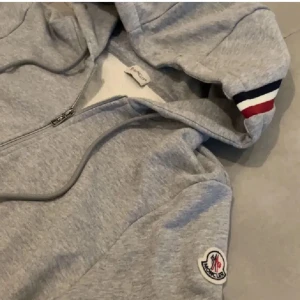 Grå hoodie från Moncler - Säljer en stilren grå hoodie från Moncler med dragkedja och broderad logga på ärmen. Tröjan har ribbade muddar med röd och blå randdetalj samt en praktisk huva med dragsko.