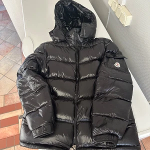 Svart pufferjacka från Moncler - Snygg svart pufferjacka från Moncler med huva och dragkedja. Jackan har en quiltad design och en ikonisk logga på ärmen. Perfekt för kalla dagar.