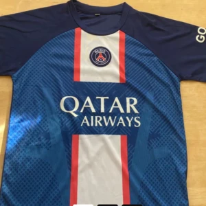 Blå PSG fotbollströja - Snygg blå PSG fotbollströja med korta ärmar och röd-vit randig detalj på framsidan. Tröjan har Qatar Airways-logga och PSG-emblem på bröstet. Perfekt för fotbollsfans!