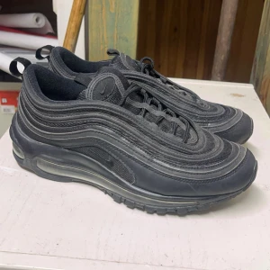 Nike Air Max 97 - Säljer ett par svarta Nike Air Max 97 med ikonisk vågig design och synlig Air-enhet i sulan. Skorna har snörning och är tillverkade i syntetmaterial för en modern look.