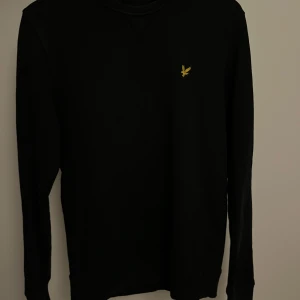 Svart långärmad tröja från Lyle & Scott - Säljer en stilren svart långärmad tröja från Lyle & Scott med ett broderat gult emblem på bröstet. Perfekt för vinter. Mycket bra skick