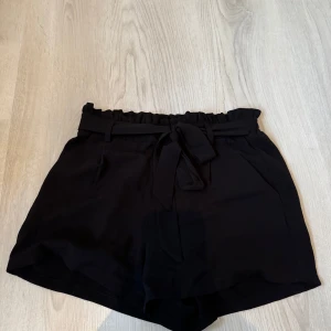 Svarta shorts med knytband - Snygga svarta shorts med hög midja och knytband i midjan. Perfekta för en stilren look under varma dagar. De har en lös passform och är tillverkade i ett mjukt material för extra komfort.